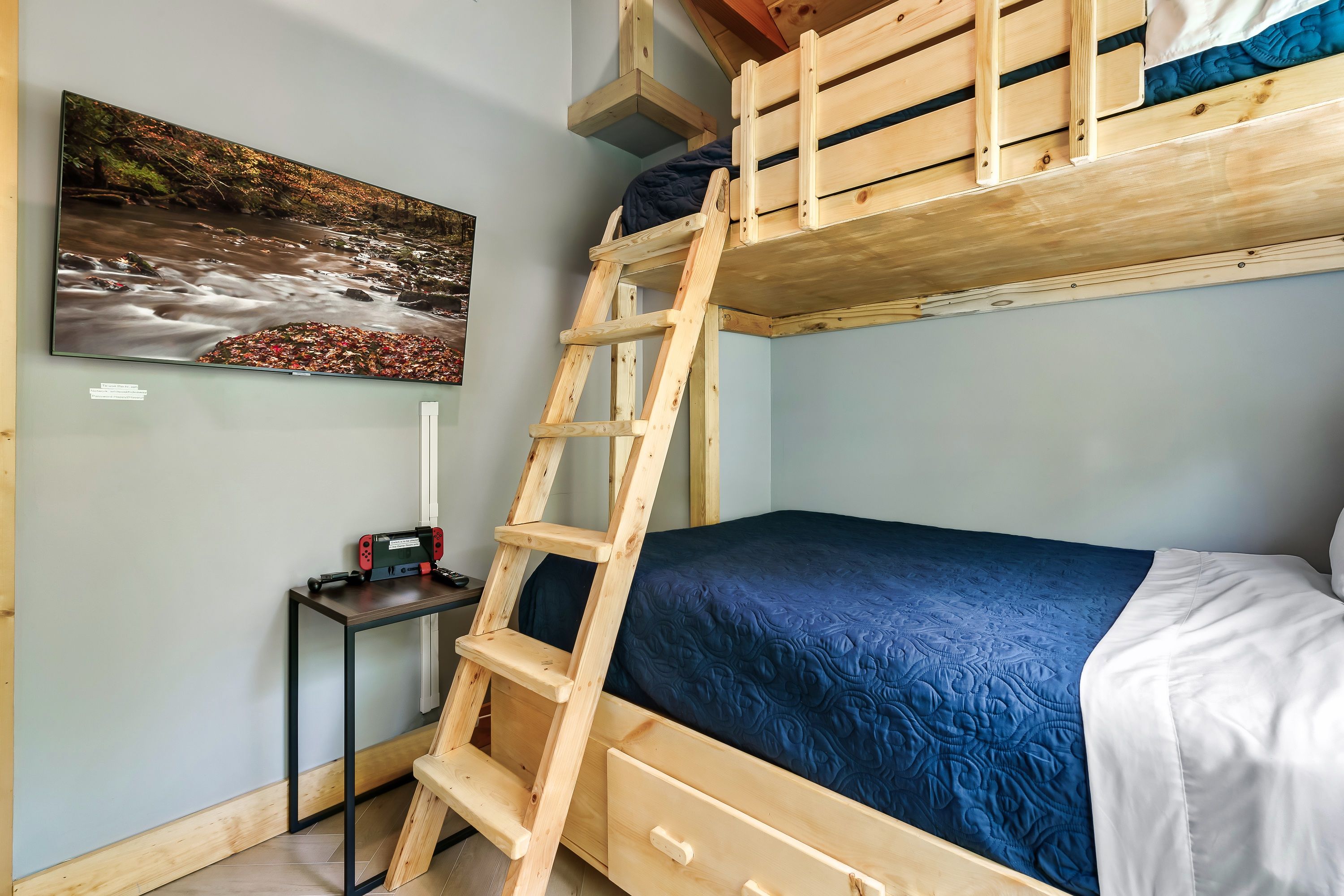A bunk bed with a wooden ladder, a desk with a chair, and a painting of a river.
