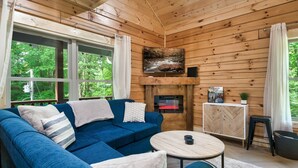 A cozy cabin interior with a blue sofa, a fireplace, and large windows offering a view of the forest. - Modern cabin w/heated pool & hot tub next to pkwy (Sevierville)