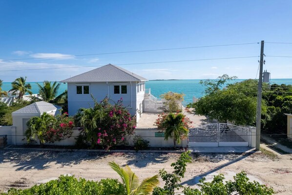 A white house with a grey roof, surrounded by lush greenery and a view of the ocean. - Ocean Front Property With Pool. - George Town (Exuma)