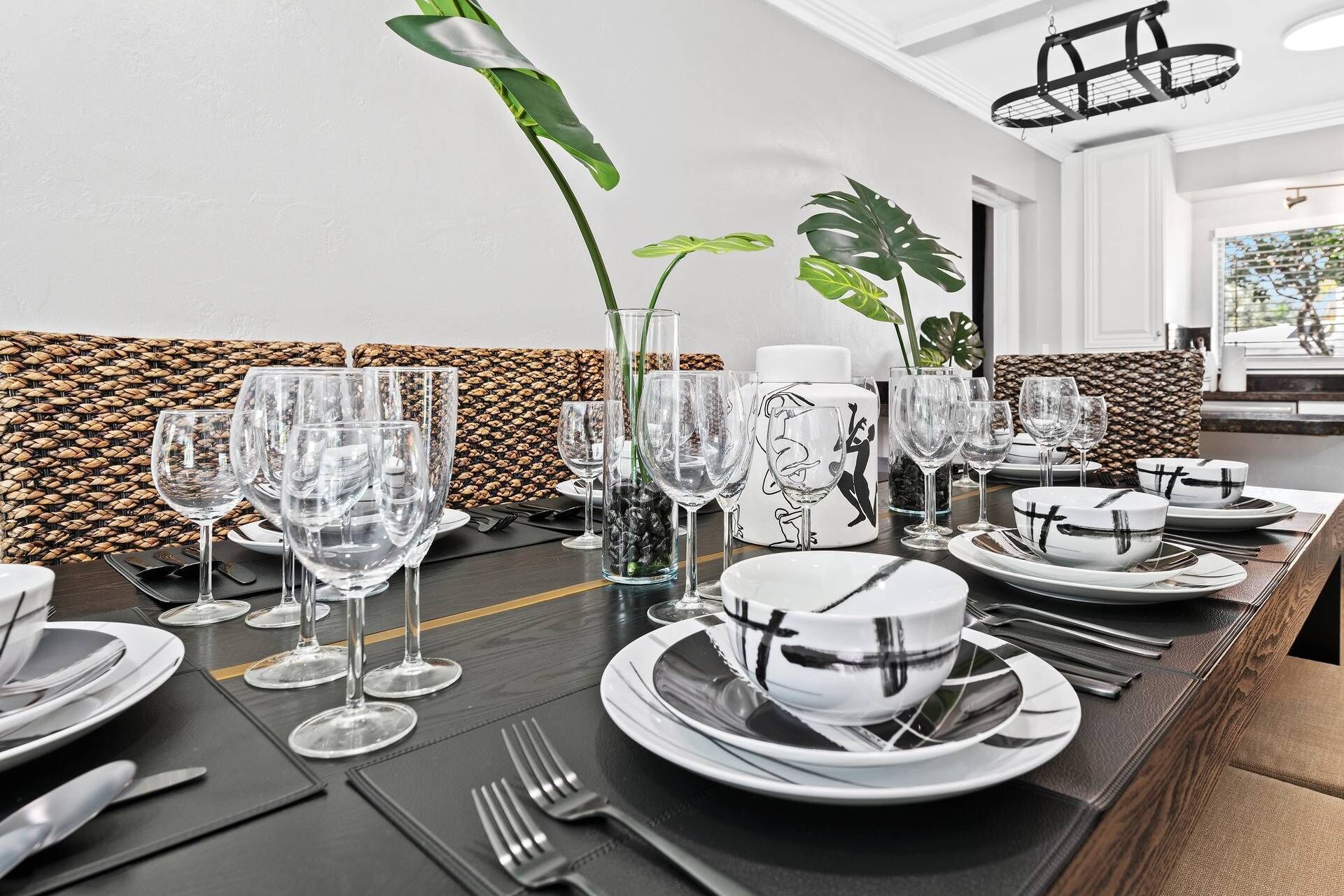 A dining table set with multiple wine glasses, plates, and cutlery, with a woven wall panel and a plant in the background.