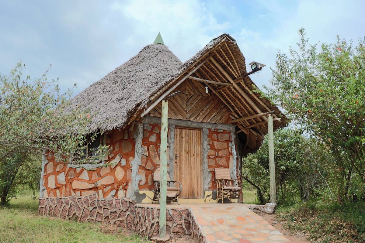 A rustic stone and thatch hut with a wooden door, a small porch, and a simple wooden table and chair set.