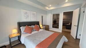 A bedroom with a large bed, bedside tables with lamps, framed artwork on the wall, and a view into the living area through an open door.