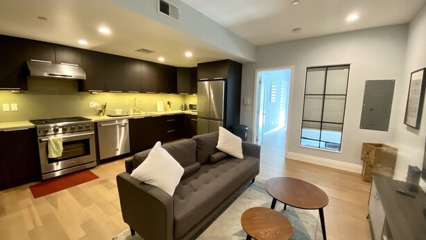 A modern kitchen with stainless steel appliances, a dark-colored sofa with white cushions, and a small wooden table.