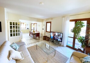 Amplia sala de estar con una mesa de centro de vidrio, un sofá y un televisor. Se observa un comedor con una mesa y sillas al fondo.
