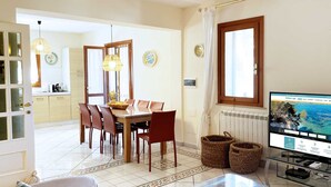 Un salon bien éclairé, comprenant un coin repas, un téléviseur et une cuisine visible au fond.