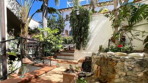 Une cour avec des marches en pierre menant à un bâtiment blanc, entouré d’une végétation luxuriante et agrémentée d’un petit élément décoratif aquatique.