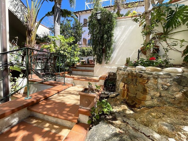 A courtyard with stone steps leading to a white building, surrounded by lush greenery and a small water feature.