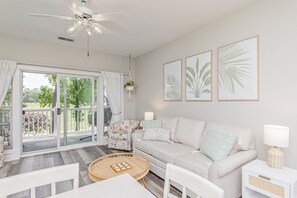 A living room with a white sofa, a round table, and a ceiling fan. There are framed pictures on the wall and a window with curtains.