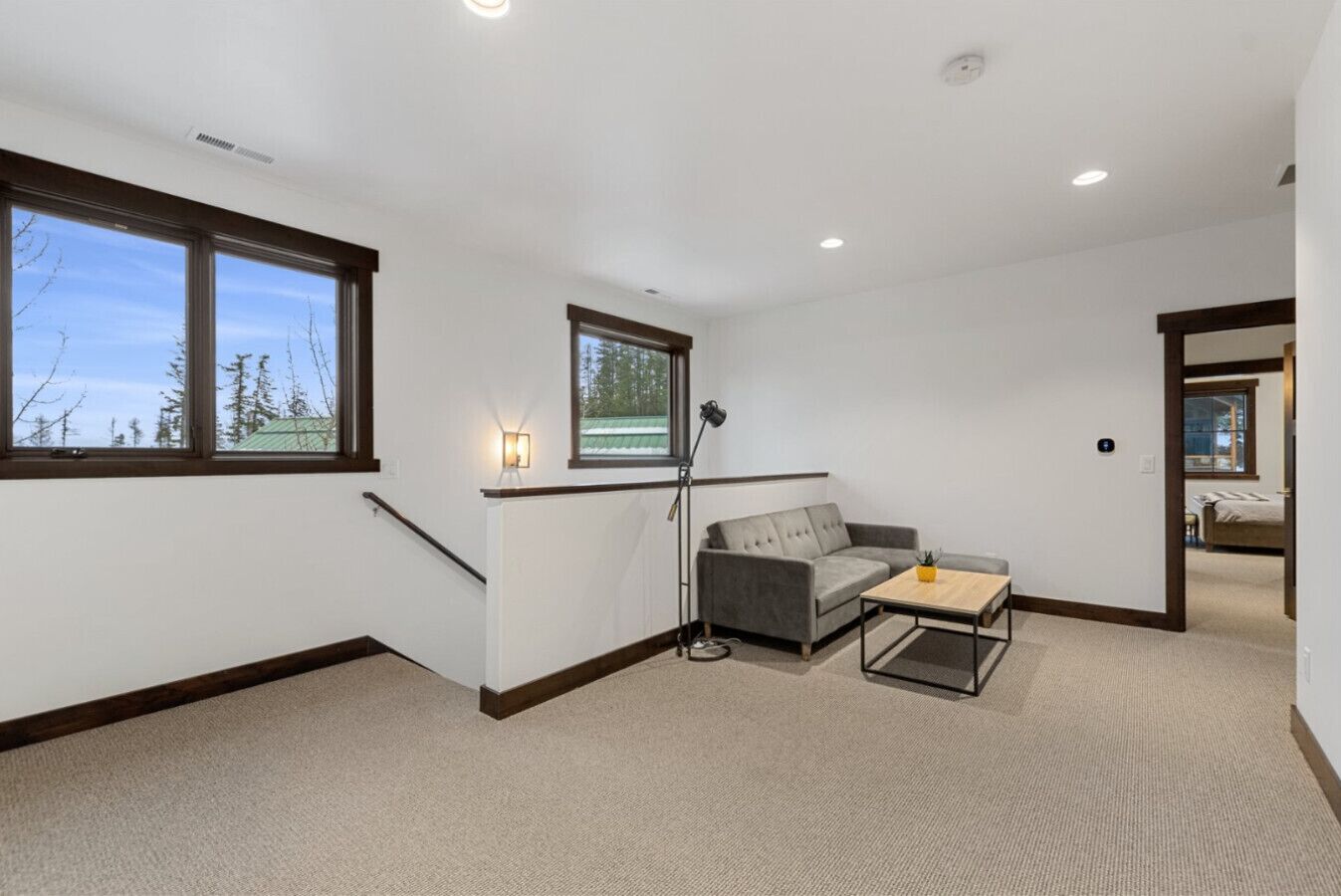 A spacious room with a grey sofa, a small wooden coffee table, and a floor lamp. There are large windows providing natural light and a view of trees.