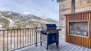 A balcony with a barbecue grill, a view of snow-covered mountains, and a wooden cabin structure.