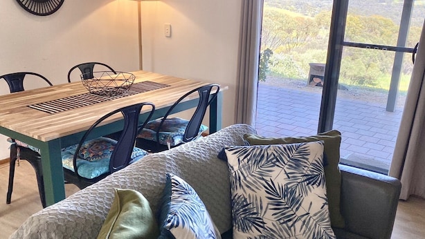 Un salon chaleureux avec une table à manger en bois, un canapé agrémenté de coussins à motifs, une lampe sur pied et une vue sur l’extérieur grâce à une grande fenêtre.