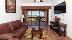 A living room with a brown leather sofa, a wooden coffee table, a ceiling fan, a TV mounted on the wall, and a view of the outdoors through a sliding glass door. - Marina Pinacate Remodeled 3 Bedroom 2 Bath Overlooking Sea of Cortez Views (Puerto Penasco)