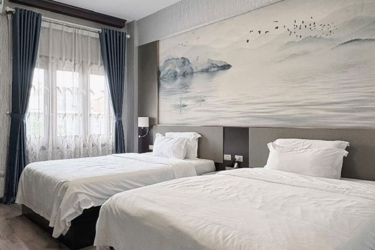 A hotel room with two beds, a large painting of a mountainous landscape, and a window with curtains.
