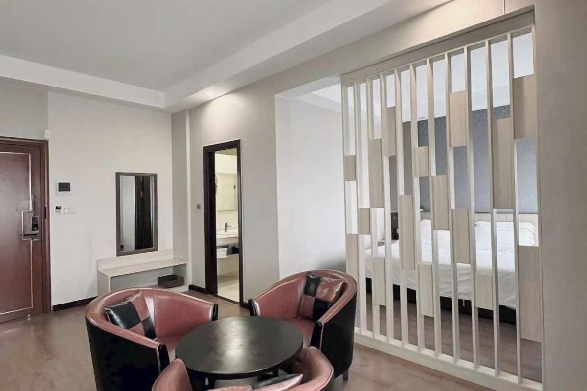 Een hotelkamer met een bed, een ronde tafel met stoelen, een spiegel en een roomdivider met verticale lamellen.