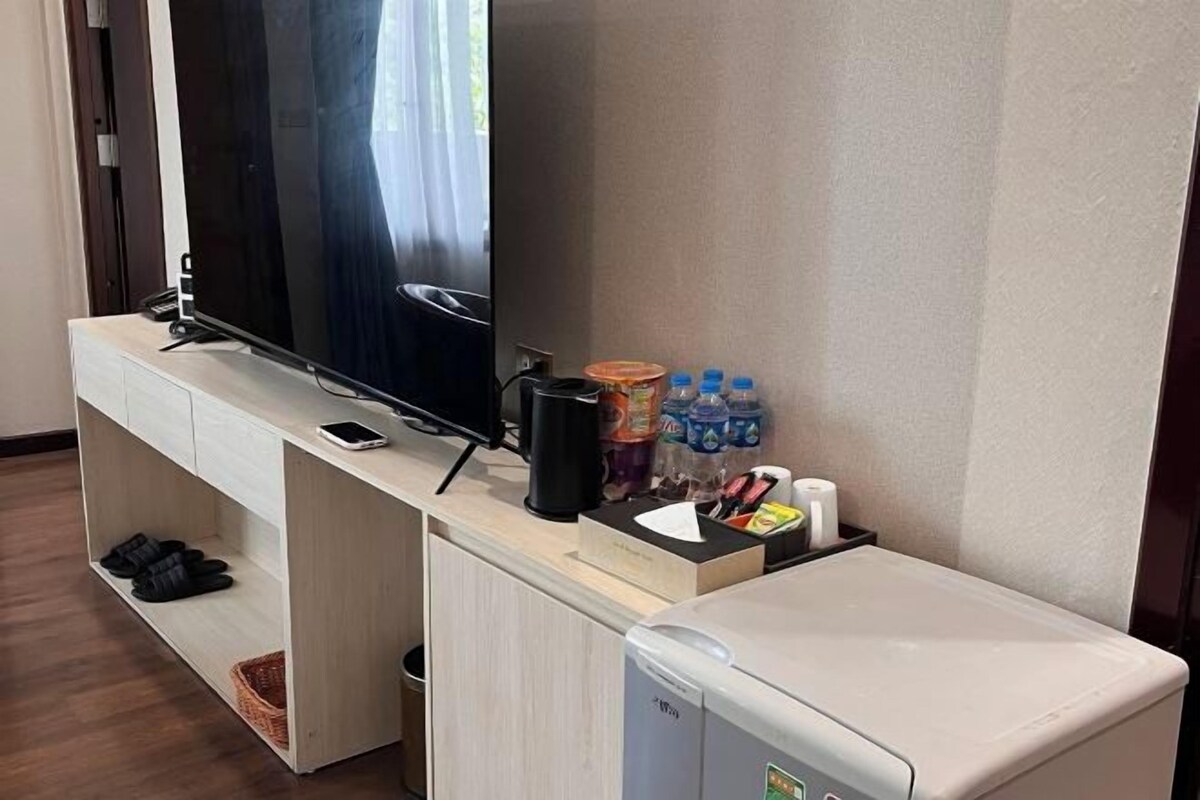 A compact hotel room with a TV, a small refrigerator, a coffee maker, and a shelf with slippers and toiletries.