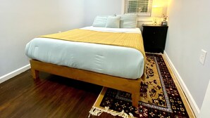 A bedroom with a bed, a nightstand, a window with blinds, and a patterned rug. - Whisper Of Himalaya (Sylva)