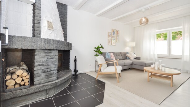 Ein modernes Wohnzimmer mit Kamin aus Stein, einem grauen Sofa, einem Holztisch und einem großen Fenster mit Vorhängen.