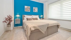 Ein modernes Schlafzimmer mit blauer Akzentwand, einem großen Bett mit beiger Kopfteil sowie zwei Nachttischen mit Lampen und einem Fenster mit Jalousien.