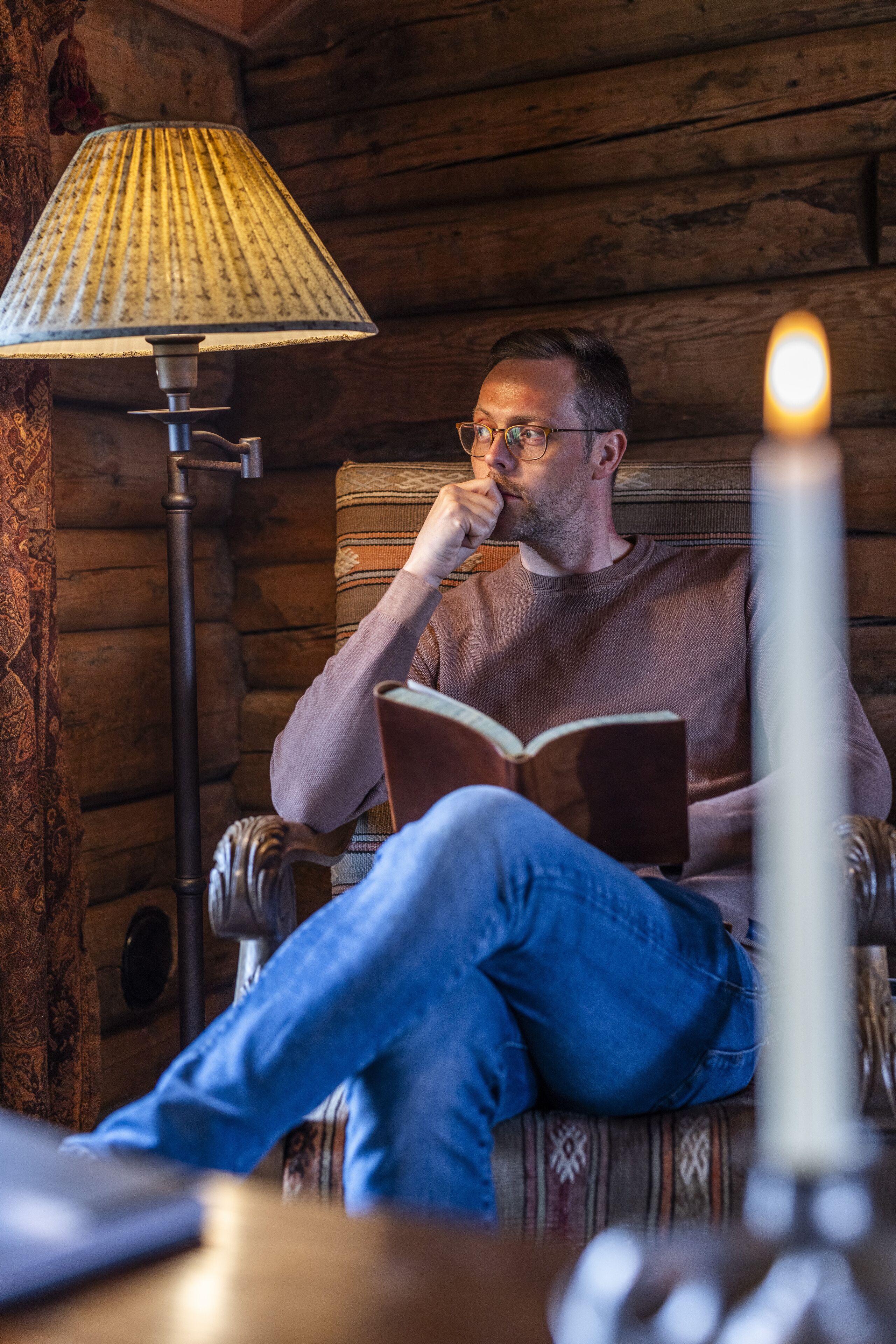 Um homem sentado em uma cadeira, lendo um livro em um quarto de cabana de madeira, com uma lâmpada e uma vela sobre uma mesa.