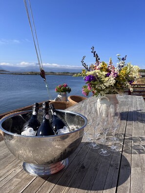 Una terraza de madera con un cubo metálico que contiene dos botellas de champán y varios vasos, un arreglo floral y vistas al agua y montañas distantes.