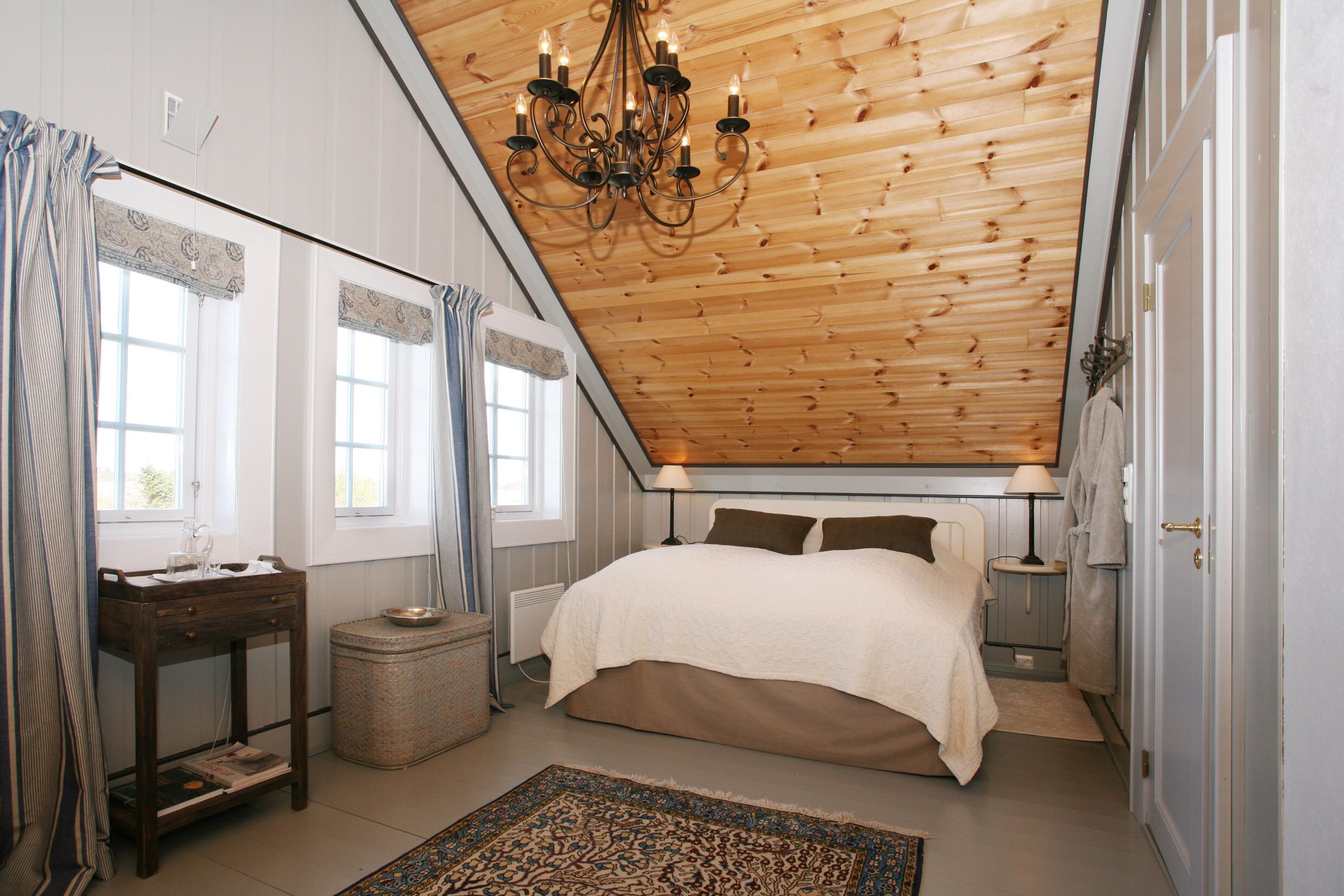 A bedroom with a sloped ceiling, a bed with a white and brown duvet, a bedside table with a lamp, and a window with curtains.