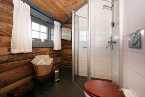 Un baño rústico en una cabaña de madera, con ducha, inodoro, ventana con cortinas y una cesta de toallas.