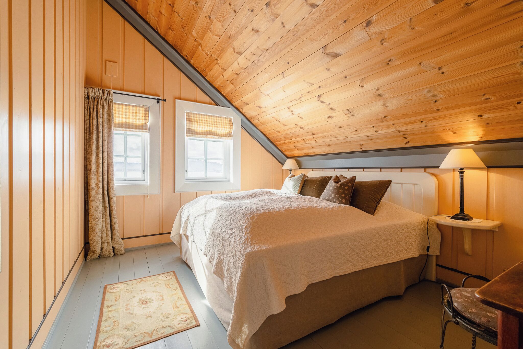 A bedroom with a wooden ceiling, a bed with a white bedspread, a nightstand with a lamp, and a window with curtains.