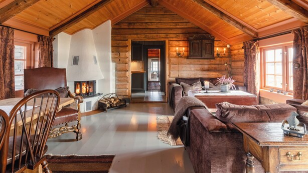 Interior de una cabaña de troncos con chimenea, mobiliario de madera y una zona de estar acogedora.