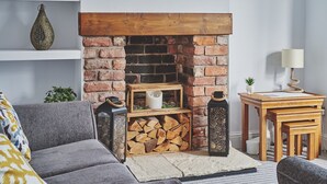 A cozy living room with a brick fireplace, a grey sofa, a small table with a lamp, and a decorative vase on a shelf. - Hollie Cottage - Luxury Cottages (Lytham Saint Annes)
