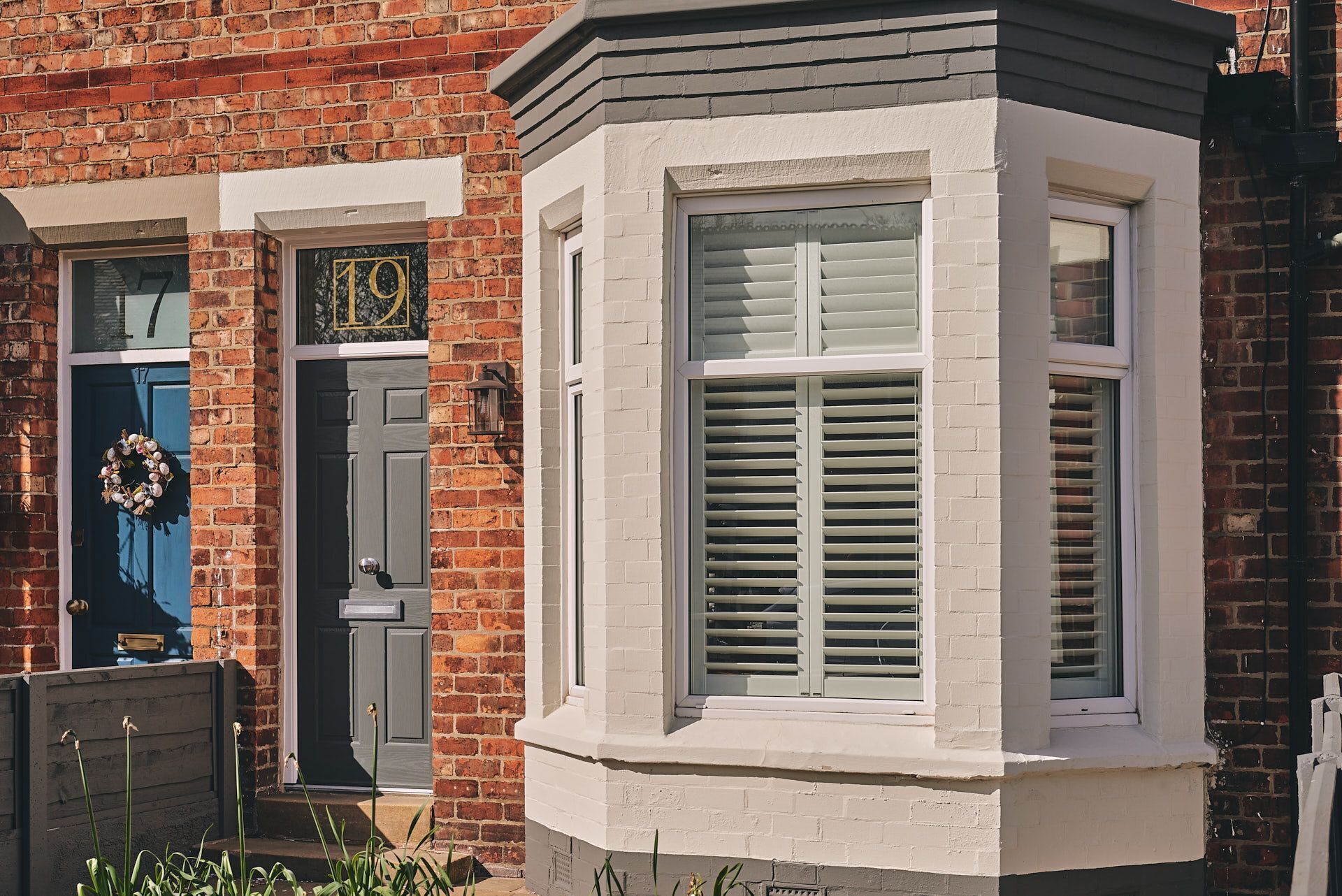 A corner bay window with white shutters, a dark blue door with a number 19 sign, and a brick wall.