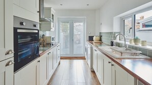 A modern kitchen with white cabinetry, a built-in oven, a sink, and a window overlooking the street. - Hollie Cottage - Luxury Cottages (Lytham Saint Annes)