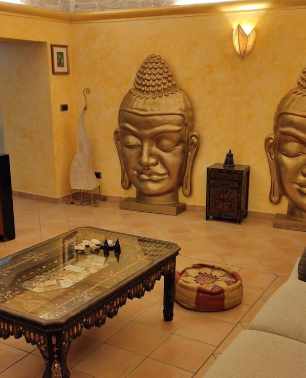 Une pièce avec une table dont le plateau est en verre, un pouf en fibres tressées et deux grandes statues de Bouddha accrochées au mur.