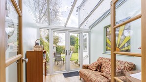 A room with a wooden floor, a patterned sofa, a wooden cabinet, and a glass door leading to an outdoor area with a table and chairs. - Bar Lane Cottage - sleeps 5 in Reepham (Reepham)