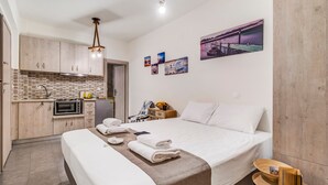 A modern hotel room with a kitchenette, a bed with neatly folded towels, a bedside table with a book and a cup, and a wall with a sunset painting. - Apartment 'Oceanida Aelia Room 2' with Shared Pool and Wi-Fi (Nikiana)