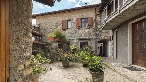 A courtyard with stone flooring, potted plants, and rustic stone buildings with wooden shutters and doors. - Casa del Picchio in Adrara San Martino (Adrara San Martino)
