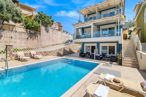 Une piscine avec des chaises longues, un mur en pierre et un bâtiment à deux étages doté de balcons.