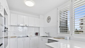 Una cocina moderna con armarios blancos, una campana extractora de acero inoxidable, horno empotrado, cafetera y una ventana con persianas.