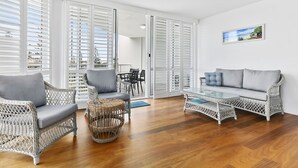 Un amplio salón con muebles de mimbre, ventilador de techo y ventanas grandes con contraventanas.