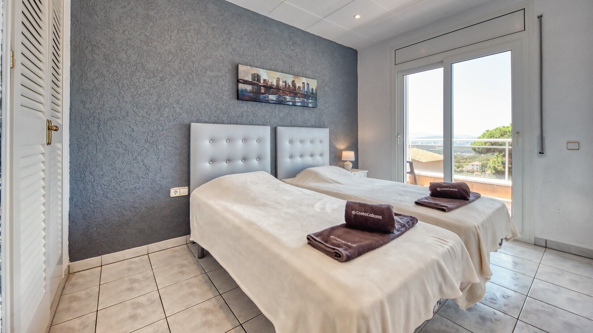 A hotel room with two beds, a cityscape painting on the wall, and a large window offering a view.