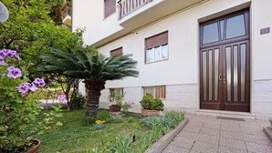 A building with a large entrance door, a small garden with various plants, and a paved walkway.
