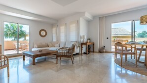A spacious living area with a sofa, coffee table, and dining table, large windows, and a view of palm trees. - Apartment 'Casa Sunchill' with Shared Pool, Wi-Fi and Air Conditioning (Marbella)