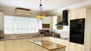 Una cocina amplia con una isla central, muebles blancos, una campana extractora negra y un gran ventanal con persianas.