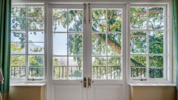 Una habitación con un ventanal grande dividido en varios paneles, una puerta blanca con pomo de latón y vistas a árboles y un balcón exterior.