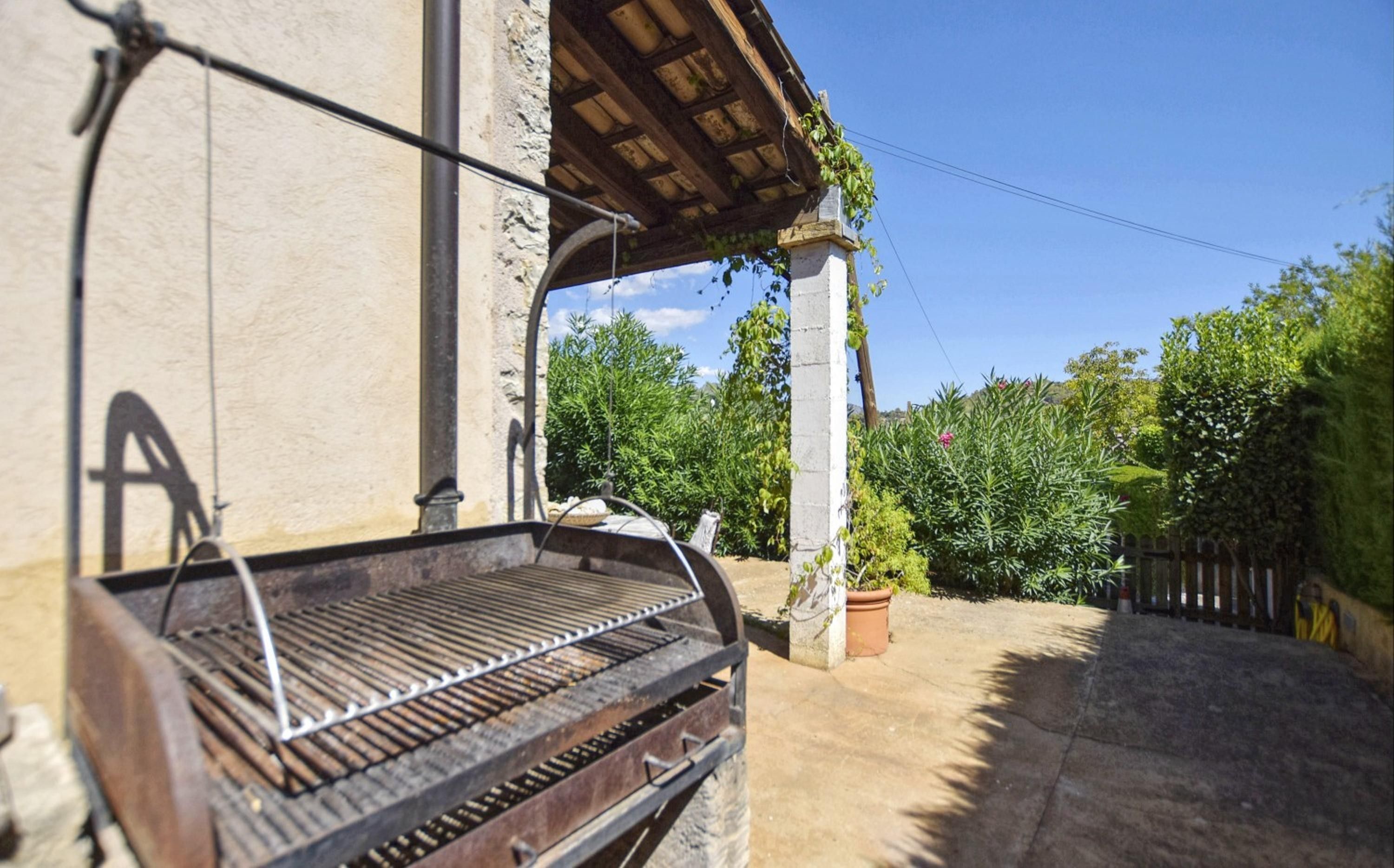 A barbecue grill outside a building with a stone facade, a wooden pergola, and a garden with various plants.