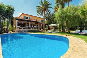 Une piscine bordée de pierres, un bâtiment avec un toit en tuiles et des palmiers.