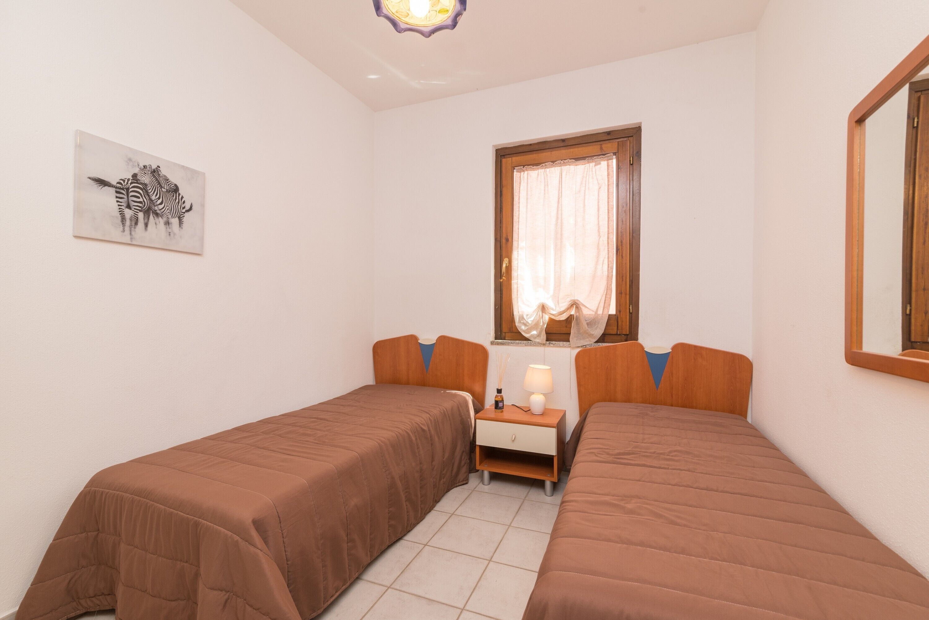 Une chambre avec deux lits, une table de chevet, une lampe, une fenêtre avec des rideaux et une œuvre d’art accrochée au mur.