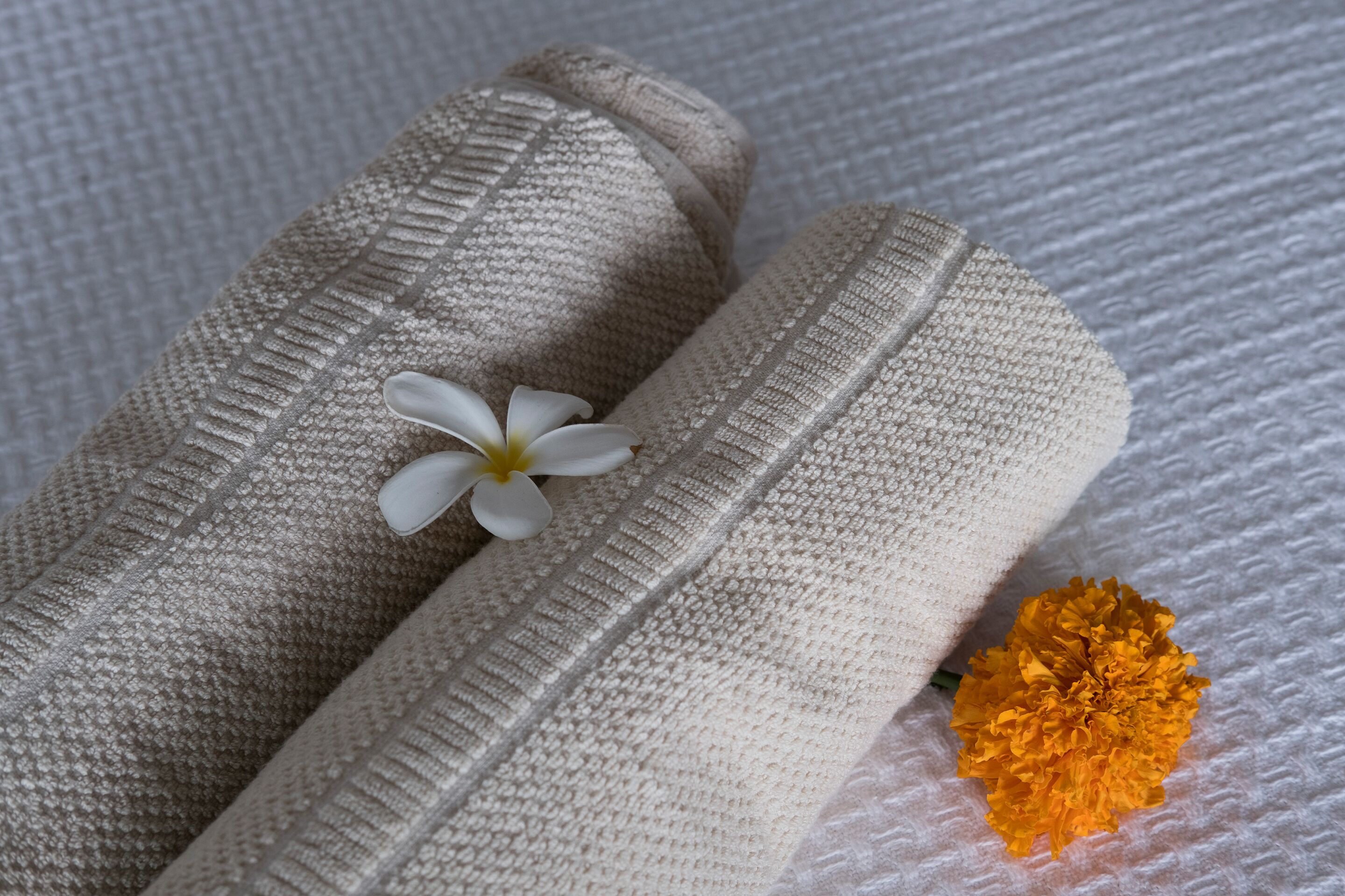 Asciugamano beige con trama, con un fiore bianco e un fiore giallo adagiati sopra.