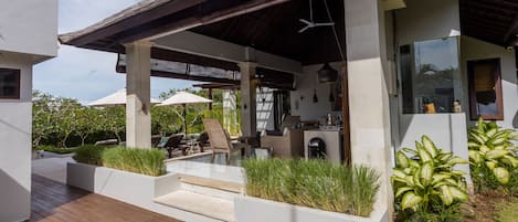 A modern outdoor patio with a wooden deck, white planters with greenery, and a covered seating area with a ceiling fan.