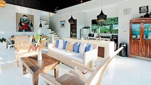 A spacious living area with a sofa, wooden coffee table, and a dining table. A kitchen with modern appliances and a view of greenery is visible in the background. - Lumi Villa Uluwatu (Pecatu)