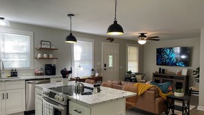 Una cocina moderna con una isla central, armarios blancos y electrodomésticos de acero inoxidable. Hay un comedor con un sofá marrón y un televisor montado en la pared.